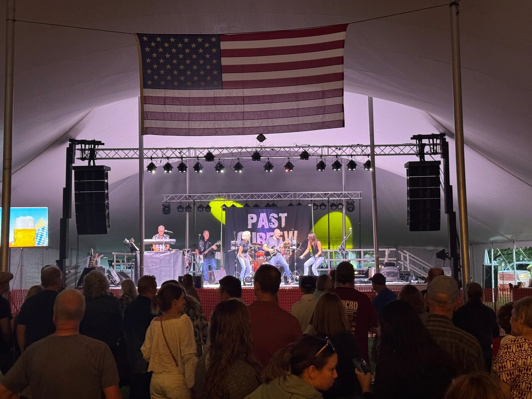 Past Curfew performs on a lit stage beneath a large American flag as a crowd watches inside a tent.