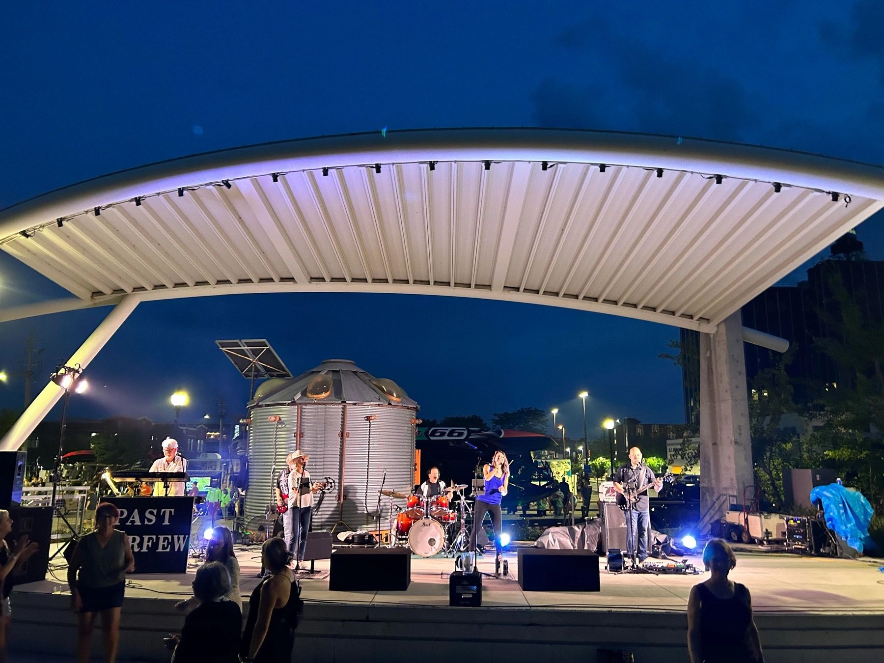Past Curfew performs with keyboards, drums and guitars beneath an outdoor canopy at dusk, with fans in front.
