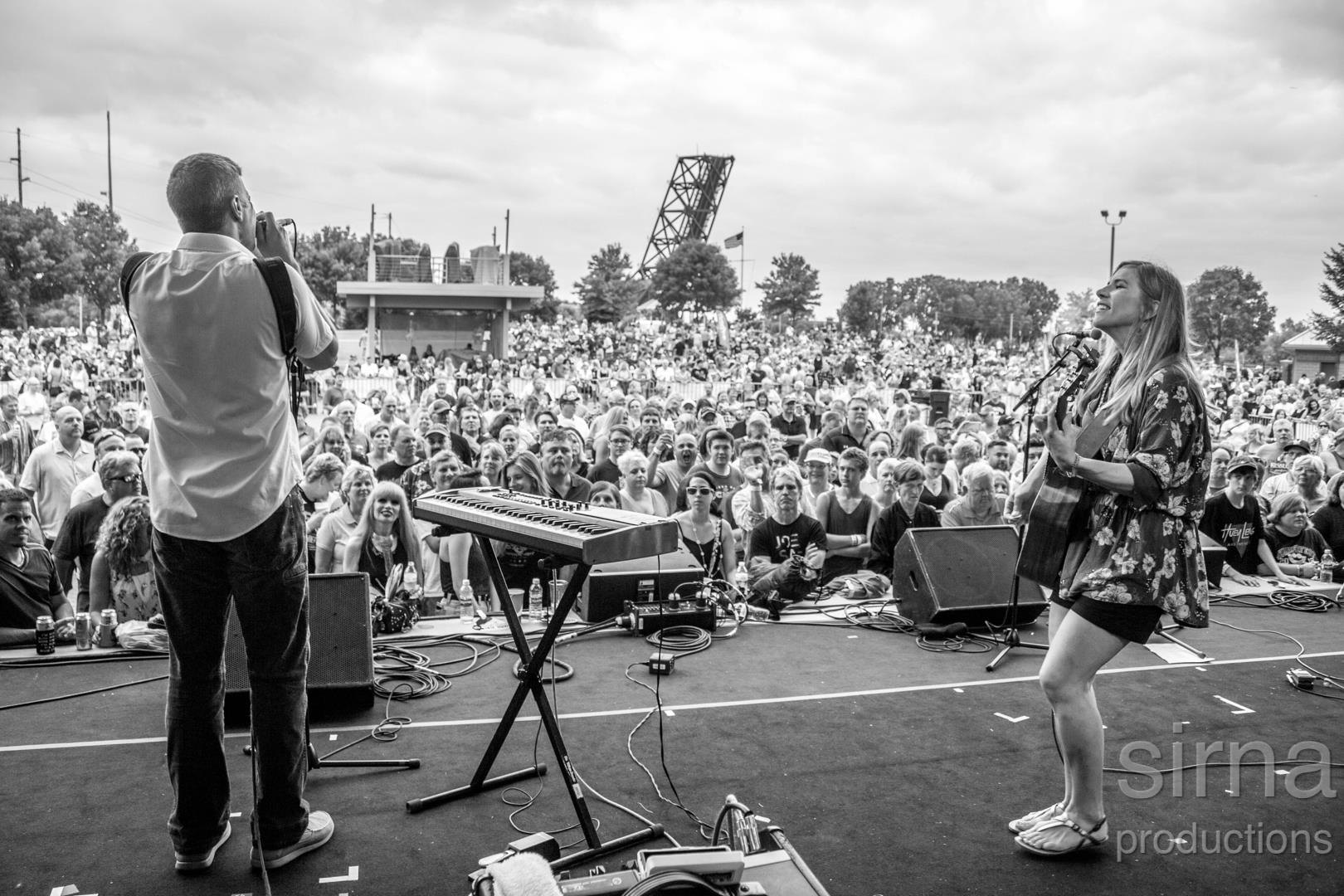 Black-and-white view of two performers with a guitar, microphone and keyboard facing a large outdoor crowd.