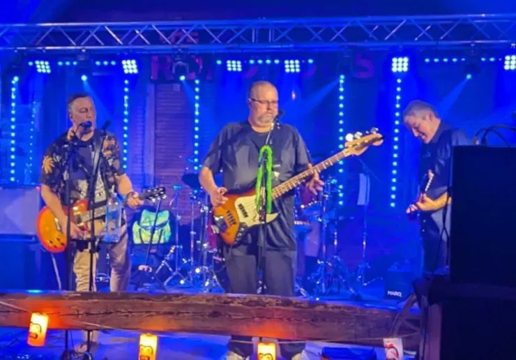 Two guitarists and a bassist perform on a blue-lit stage with microphones and a drum kit behind them.