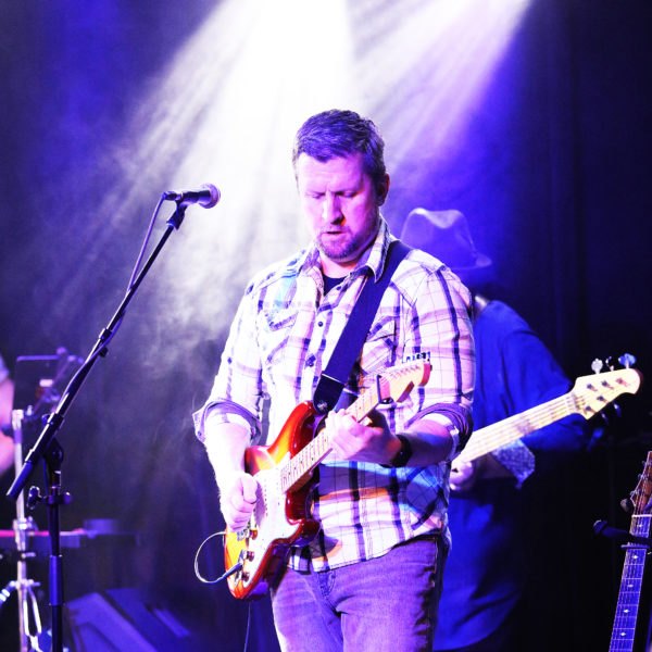 A guitarist in a plaid shirt plays under purple and white stage lights, with a bassist behind him.