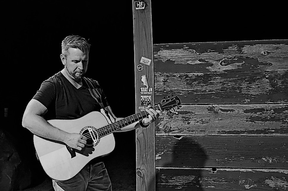 A man plays an acoustic guitar beside a weathered wooden wall in black and white.