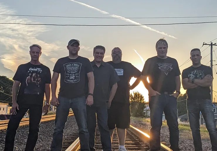 Six men in dark shirts stand together on railroad tracks, backlit by the setting sun.