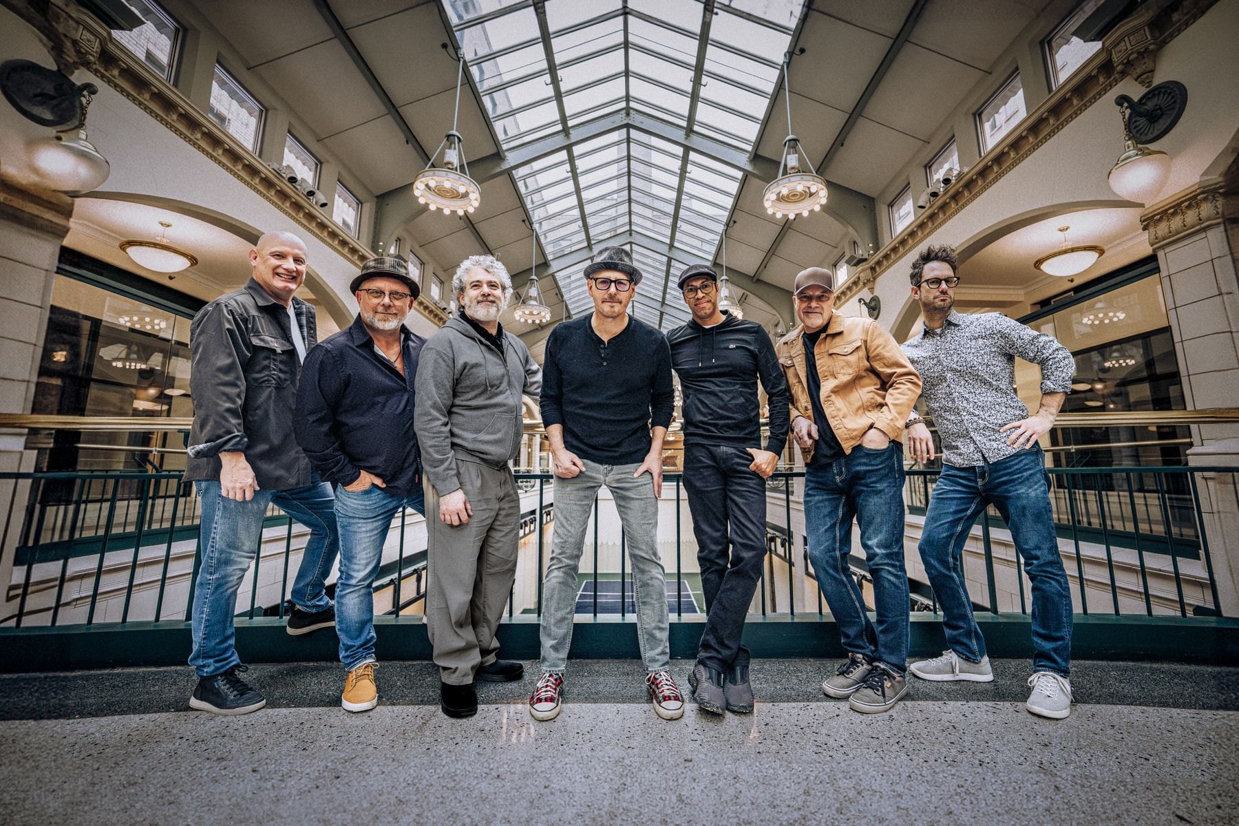 Seven men pose casually beside a balcony railing in an ornate atrium beneath a glass skylight.