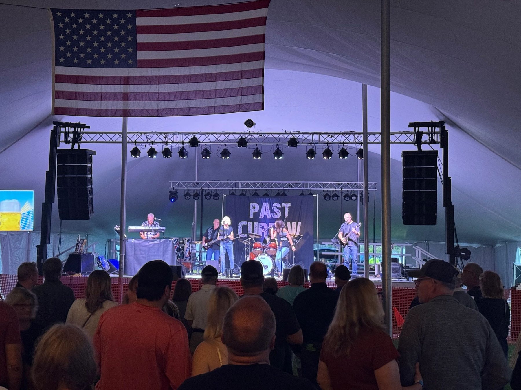 Past Curfew performs with guitars, keyboards and drums for a crowd under a tent with a large American flag.