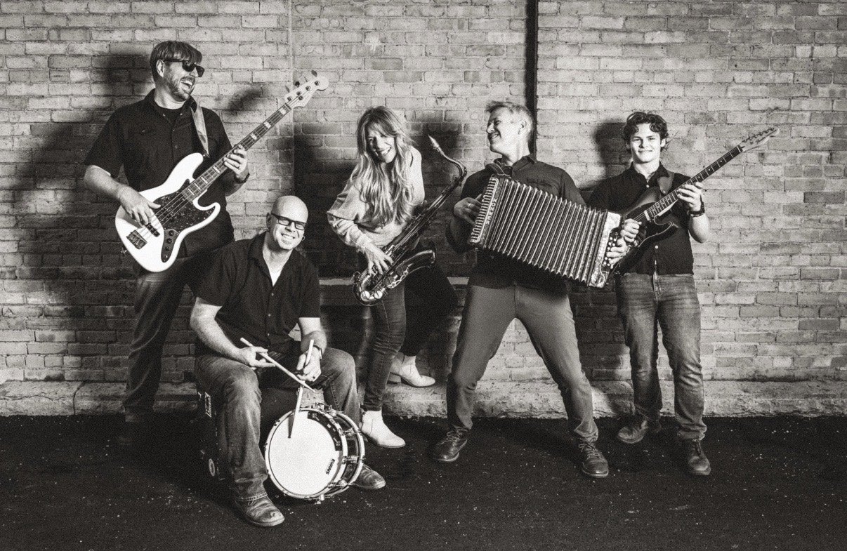 Five smiling musicians pose against a brick wall with a bass, drum, saxophone, accordion and guitar in black and white.